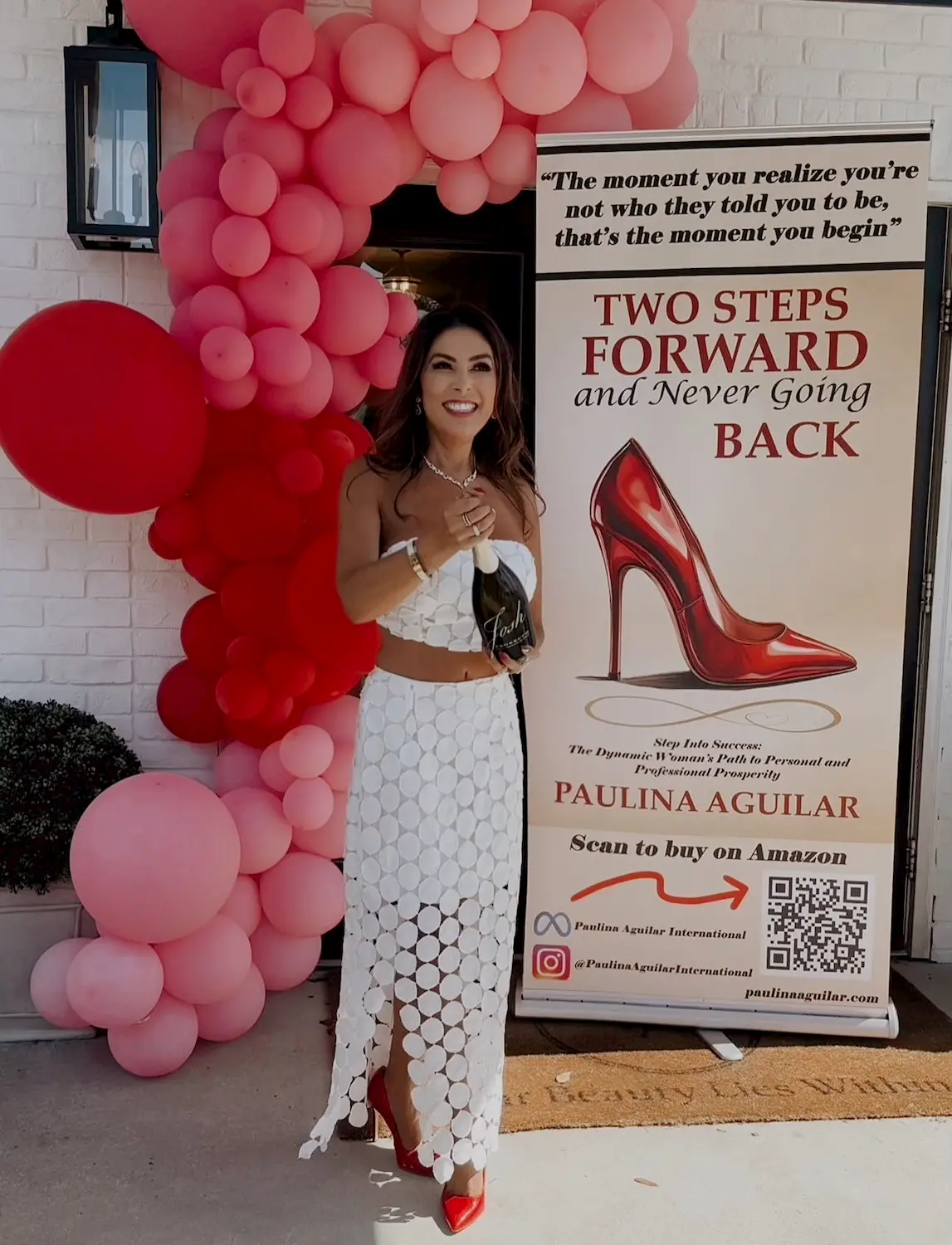 A smiling woman in a white skirt and crop top stands by a red and pink balloon arch next to a sign promoting the book "Two Steps Forward and Never Going Back" by Paulina Aguilar, featuring red high heels.