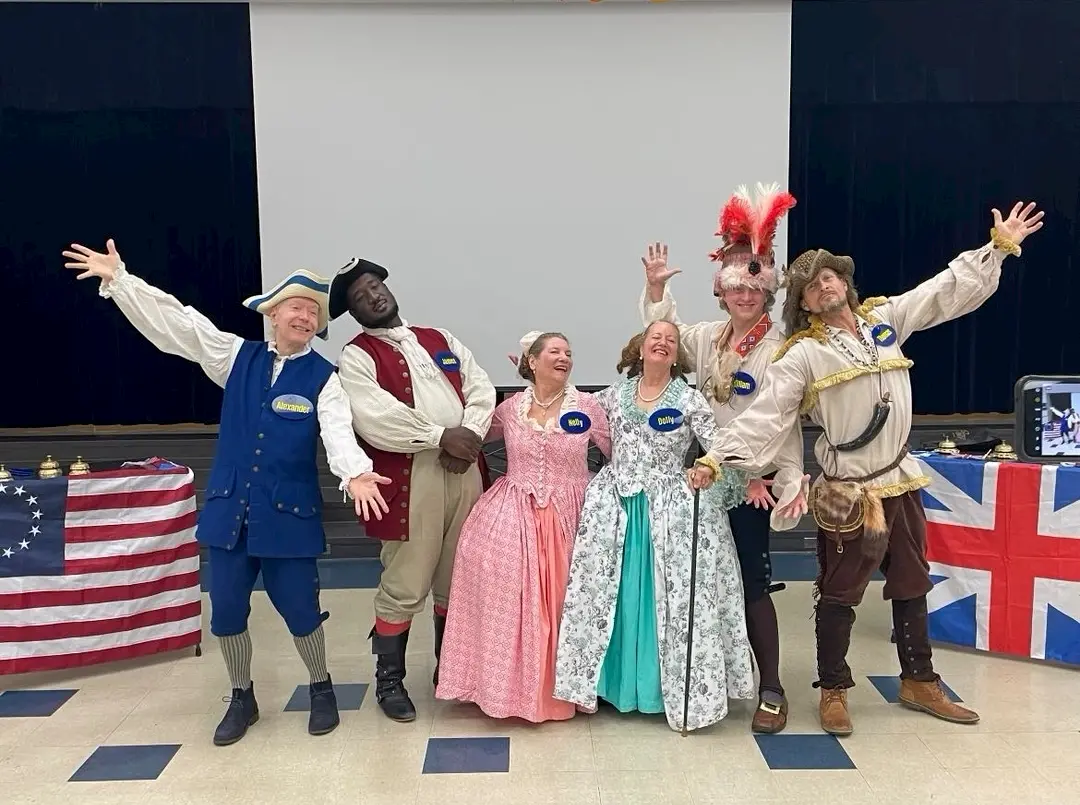 Five adults in colonial-era costumes pose joyfully, celebrating the American Revolution with arms raised, standing before U.S. and British flags. Each wears a blue badge and smiles enthusiastically at this spirited TWAM event.