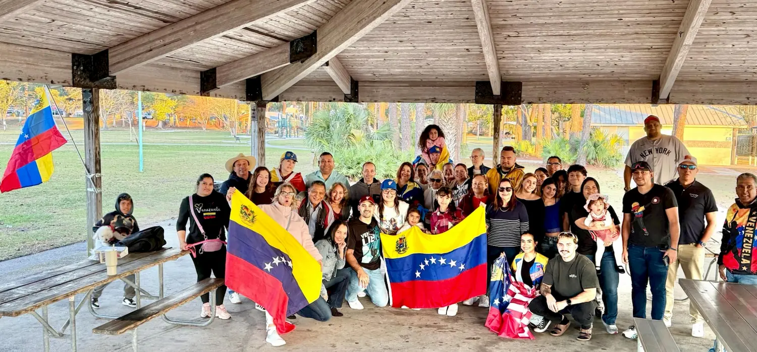 A large group of people, including adults and children, pose together under a pavilion in a park, holding Venezuelan flags and smiling at the camera. Some people are wearing festive clothing, and the background shows autumn trees.