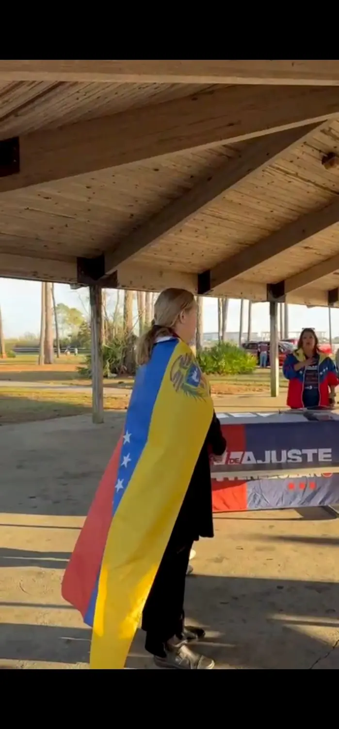 A person stands under a wooden pavilion, draped in a Venezuelan flag, facing another person near a table with a red and blue banner. Trees and parked cars are visible in the background.