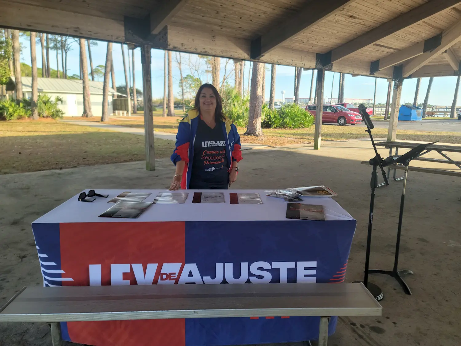 A woman stands behind a table covered with a "LEV DE AJUSTE" banner, displaying pamphlets and materials at an outdoor pavilion. Trees, cars, and buildings are visible in the background.