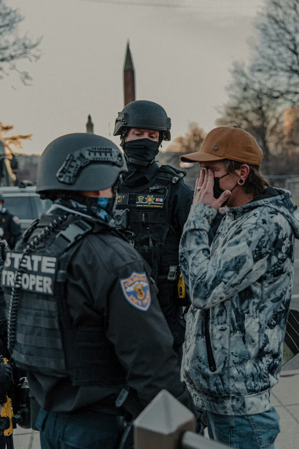 A distressed person in a camouflage jacket and brown cap covers their face while talking to two state troopers in tactical gear and helmets outdoors at dusk, after being apprehended near the US borders.