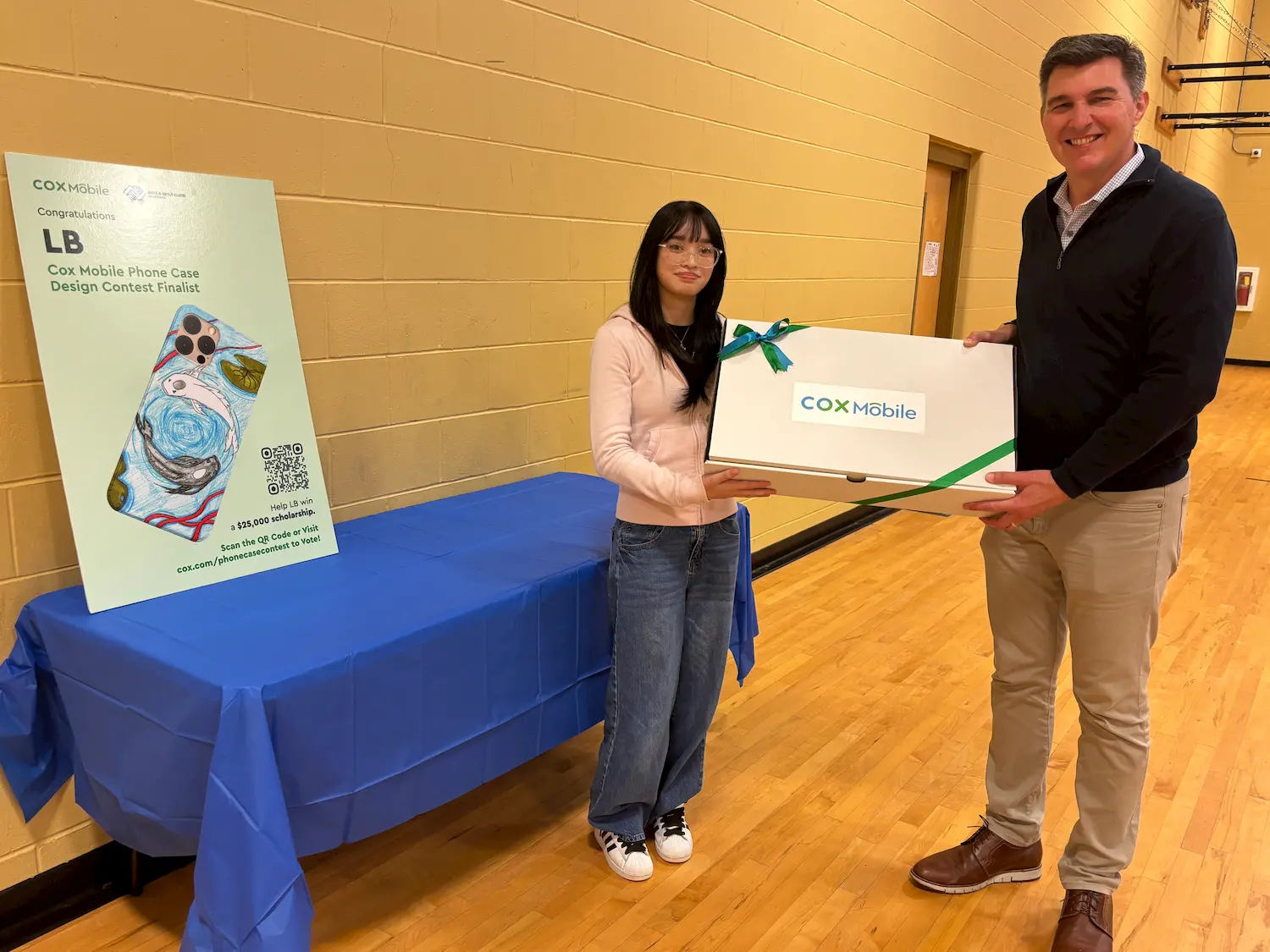 A young woman and a man stand in a gym, smiling as they hold a large white Cox Mobile box. Next to them, a table displays a poster showing a colorful phone case design and contest finalist details.