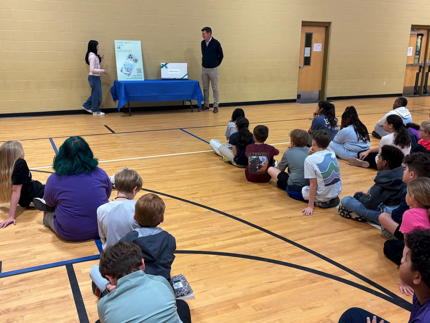 A group of children sit on a gym floor, facing a woman and a man standing by a table with a display board. The adults are presenting information, and the children are attentively listening.