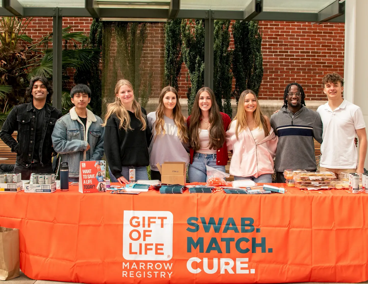 Eight smiling young adults stand behind a table with an orange "Gift of Life Marrow Registry" banner, featuring the words "Swab. Match. Cure." The table holds informational materials and snacks. Brick wall and greenery in the background.