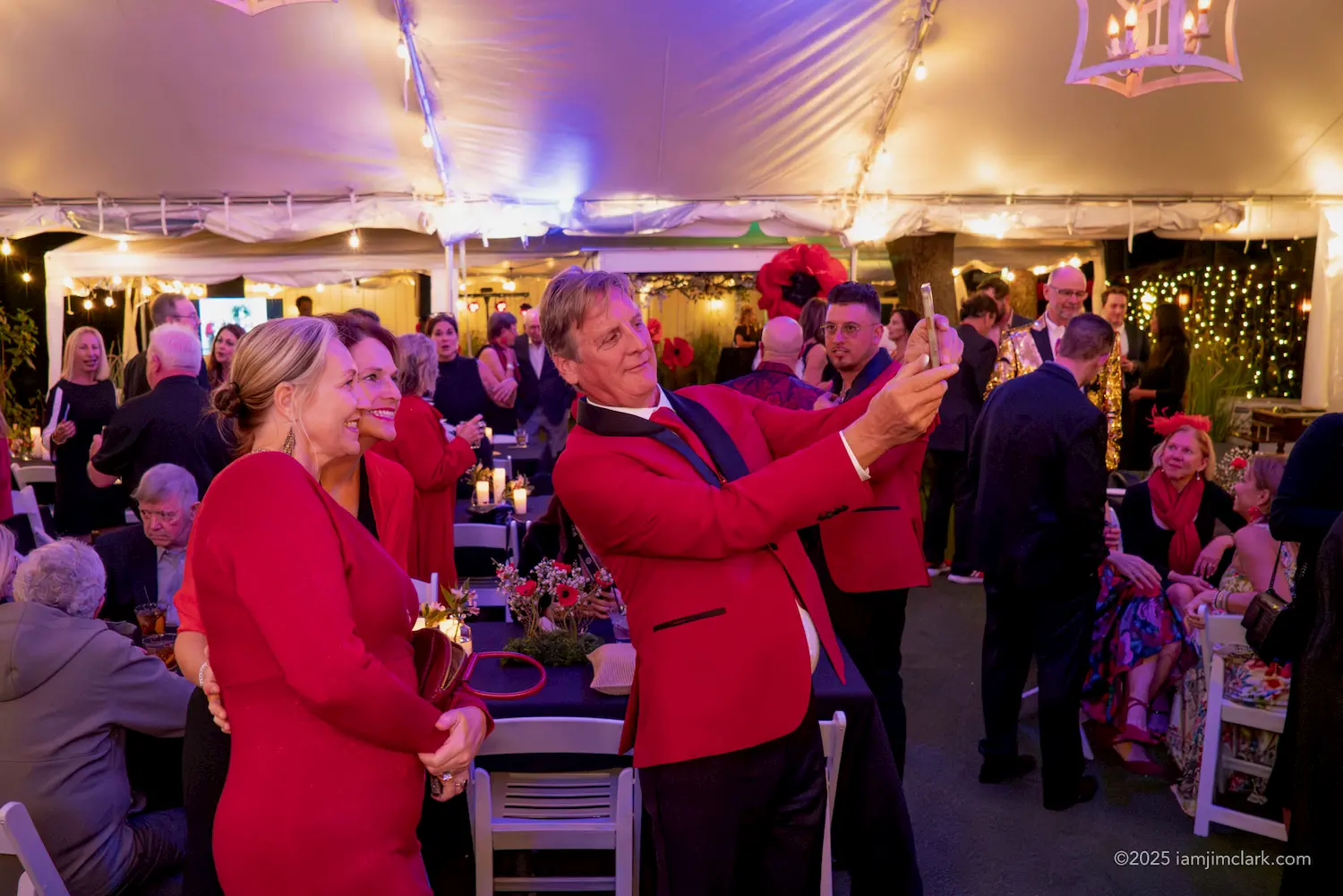 A man and woman in matching red jackets take a selfie at a lively indoor event. Guests in formal attire mingle in the festive, well-lit venue decorated with lights and holiday centerpieces.