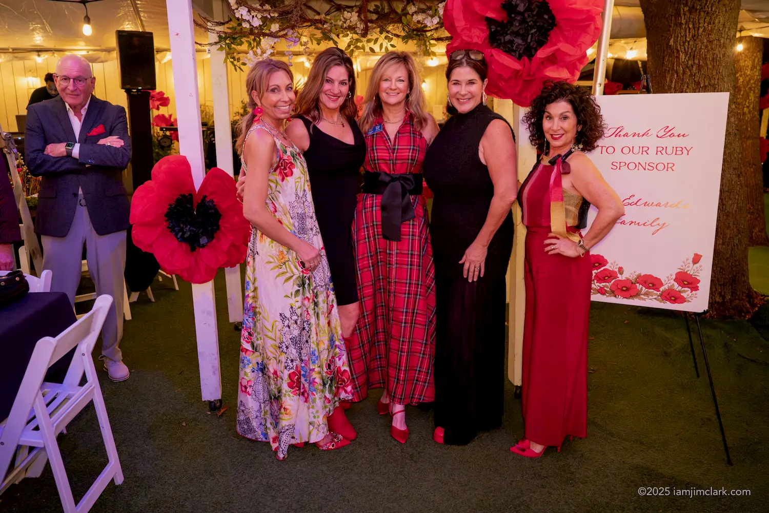Five women dressed in colorful formal attire pose and smile together at an event, while a man stands nearby. Large red flower decorations and a sponsor sign are visible in the background.