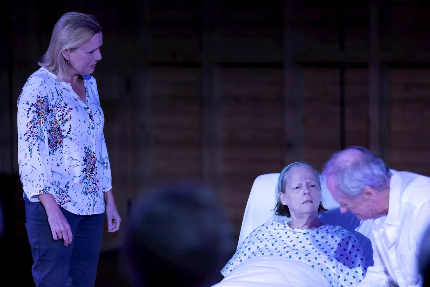 A woman in a floral blouse stands next to a bedridden elderly woman in a hospital gown, while an older man leans close to the patient, appearing concerned. The scene is dimly lit, suggesting seriousness or sadness.