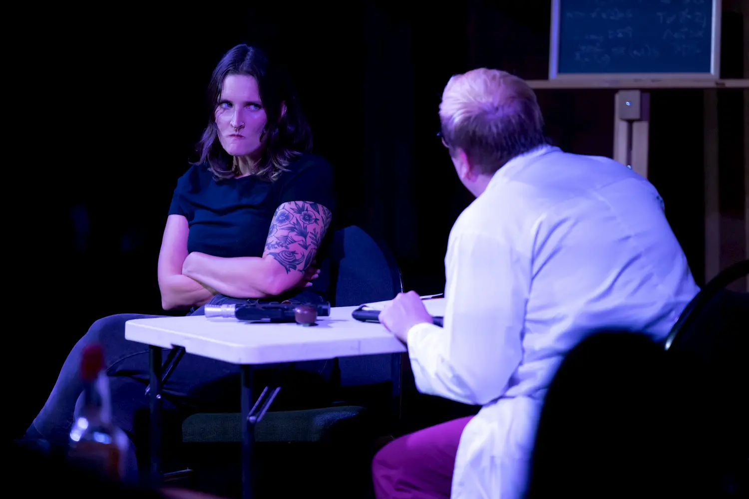 A woman with a tattoo sits at a table, arms crossed and looking upset, while a person in a white lab coat sits across from her, facing away. The setting appears tense and dramatic.