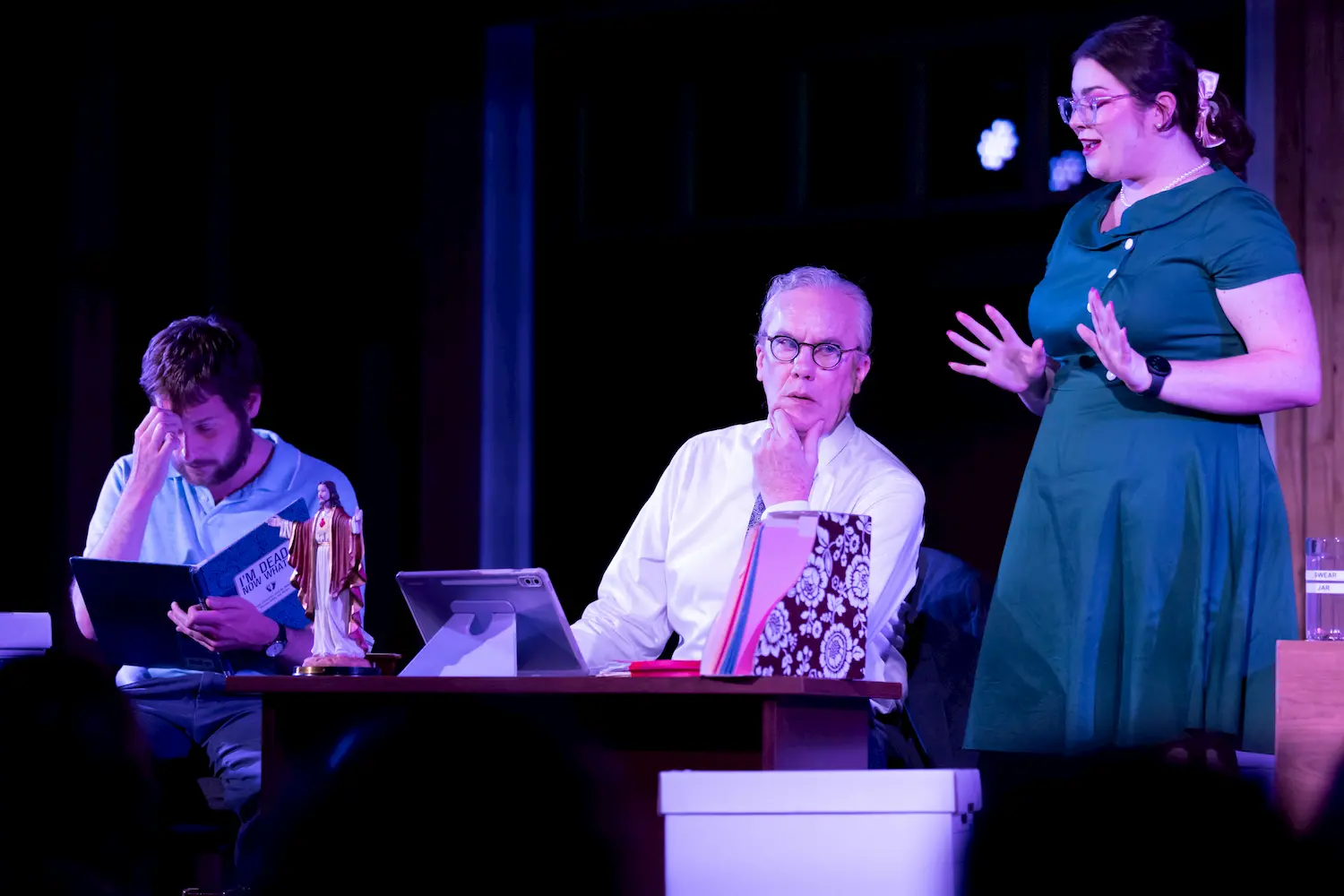 Three people are on stage in an office setting; a man with a hand to his face, another older man sitting at a desk looking thoughtful, and a woman in a green dress speaking with expressive gestures.