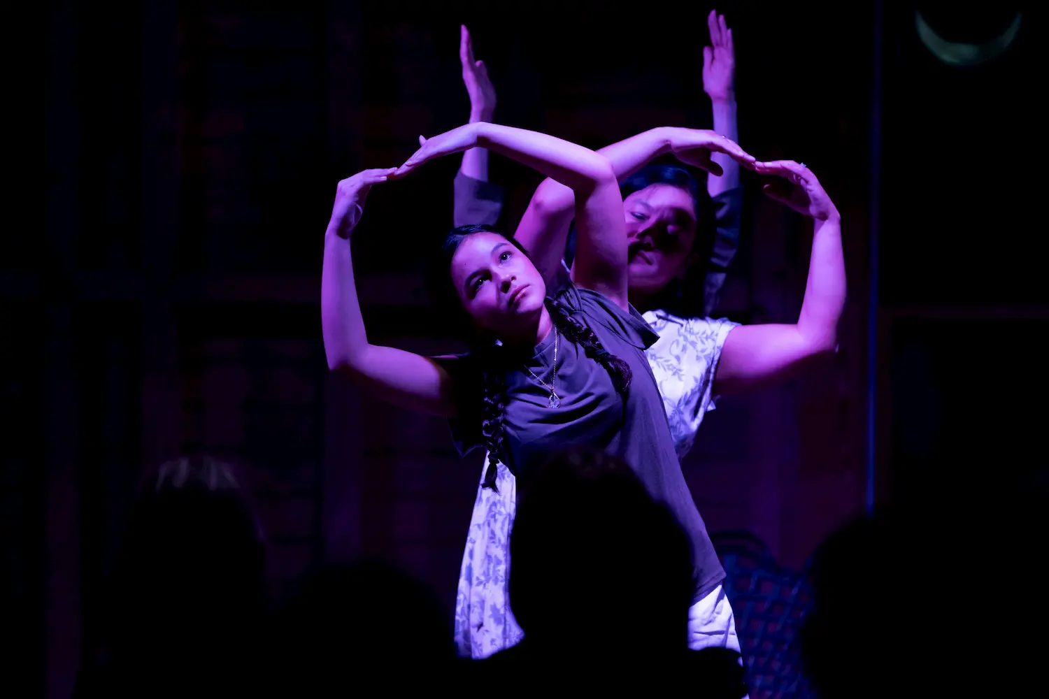 Two people perform on stage under purple lighting, standing close together with their arms raised and curved to form symmetrical shapes. The audience is visible in silhouette in the foreground.