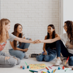 Four young women sit on the floor in a bright room, smiling and fist bumping. Art supplies and colorful drawings are spread out in front of them, suggesting a creative activity or project.