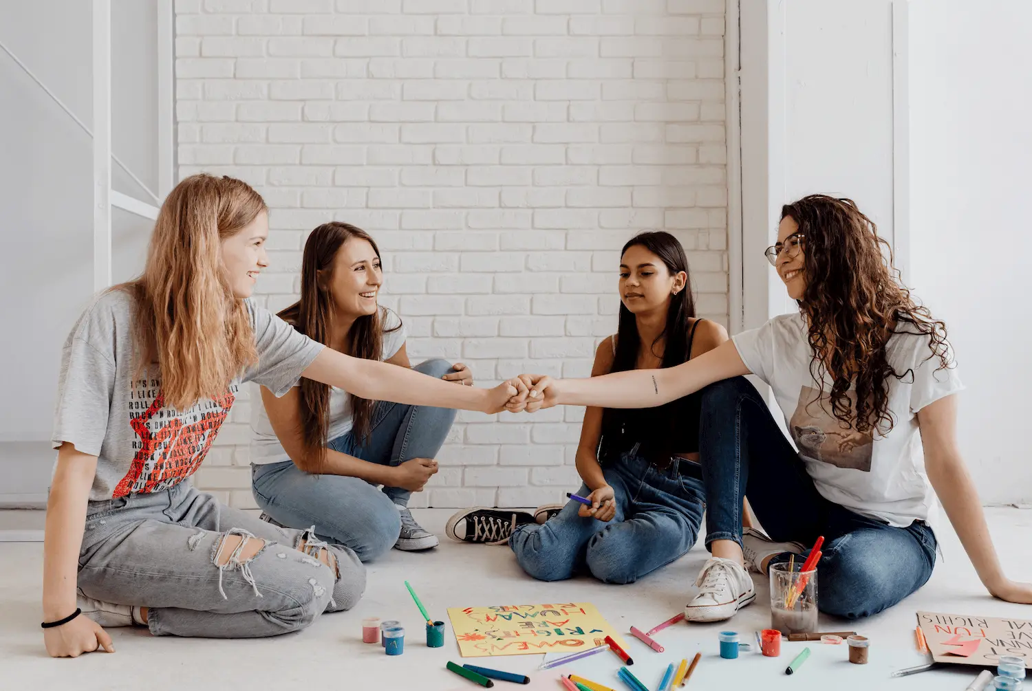 Four young women sit on the floor in a bright room, smiling and fist bumping. Art supplies and colorful drawings are spread out in front of them, suggesting a creative activity or project.