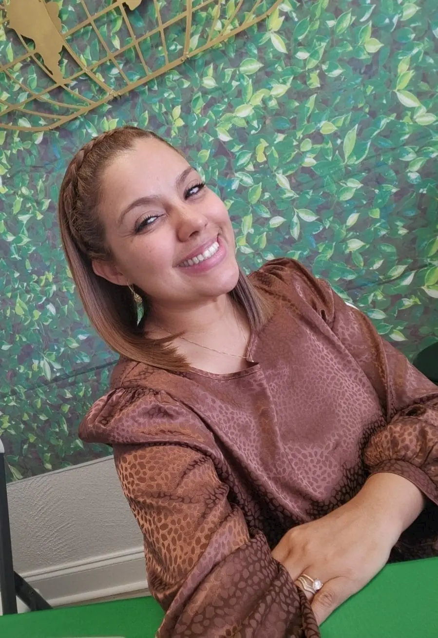 A woman with straight, light brown hair and a braided headband smiles while sitting at a table. She is wearing a brown patterned top, and the background features a green leafy design.