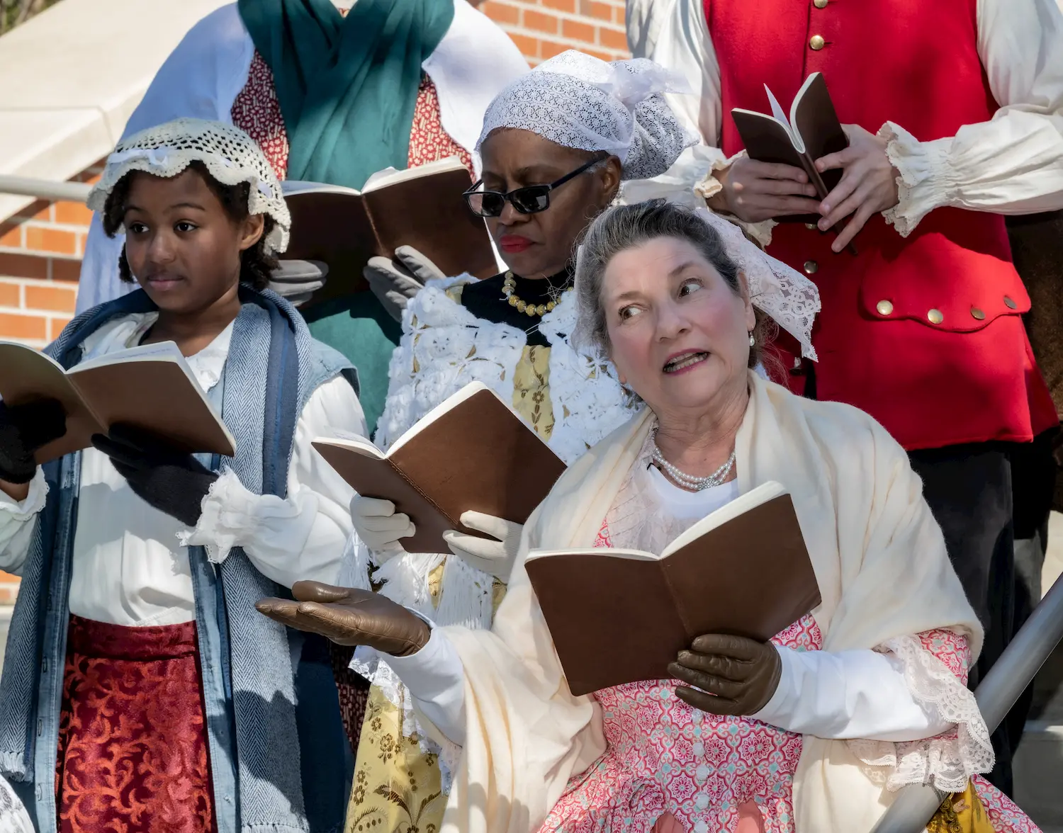 A group of people in historical clothing sing from books outdoors, with one woman in the front gesturing as she sings. They appear to be participating in a reenactment or costumed event.