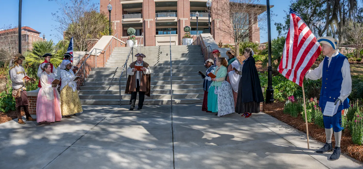 People in historical colonial costumes stand in two groups facing each other outside a brick building; one person holds an American flag, and another is reading from a document at the center of the scene near the stairs.