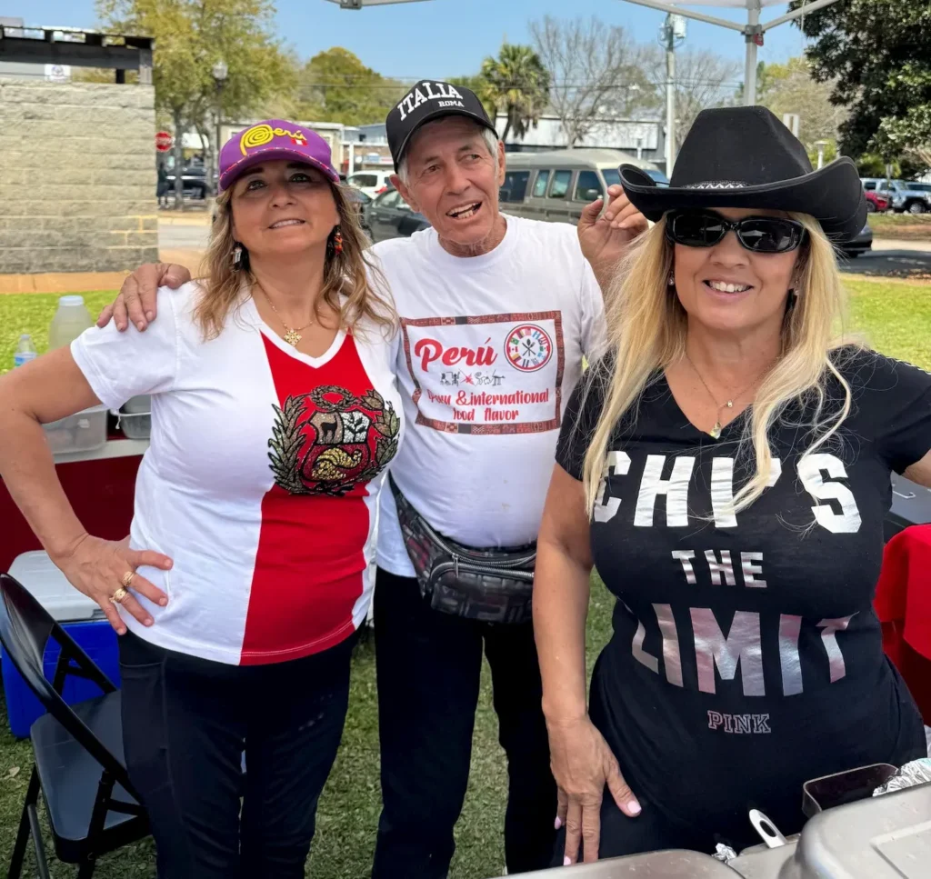 Three people pose and smile outdoors under a tent. Two women stand on either side of a man; one wears a black hat and sunglasses, the other a purple cap, and the man wears a white T-shirt and baseball cap. Trees and cars are visible behind them.