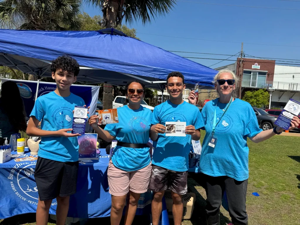 Five people wearing matching blue t-shirts stand behind a table with brochures and informational materials at an outdoor event. They are smiling and posing for the photo under a blue canopy.