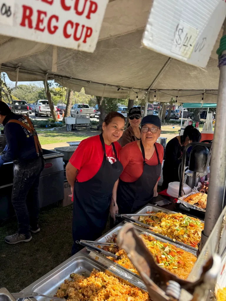 Three people wearing aprons and hats smile while standing behind trays of food at an outdoor food stall under a tent. Signs and other food items are visible in the foreground.