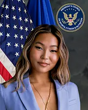 A woman with wavy, shoulder-length hair wears a light blue blazer and gold necklace, standing with coraje before an American flag and a seal reading "President's Council on Sports, Fitness & Nutrition.