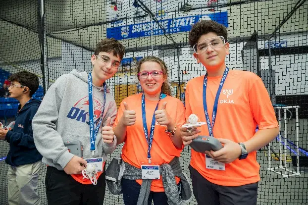 Three teens wearing safety glasses and event badges pose at a robotics competition, with two giving thumbs up and one holding a controller. They stand inside a netted area with competition equipment visible.