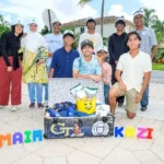 A group of nine smiling people pose outdoors around a decorated trunk filled with gifts. Colorful letters on the ground spell "UMAIR" and "KAZI." Palm trees and houses are visible in the background.