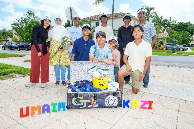 A group of nine smiling people pose outdoors around a decorated trunk filled with gifts. Colorful letters on the ground spell "UMAIR" and "KAZI." Palm trees and houses are visible in the background.