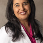 A woman with long dark hair, wearing a white blazer and a pink blouse, smiles at the camera in front of a neutral background—an advocate for raising confident kids through financial education.