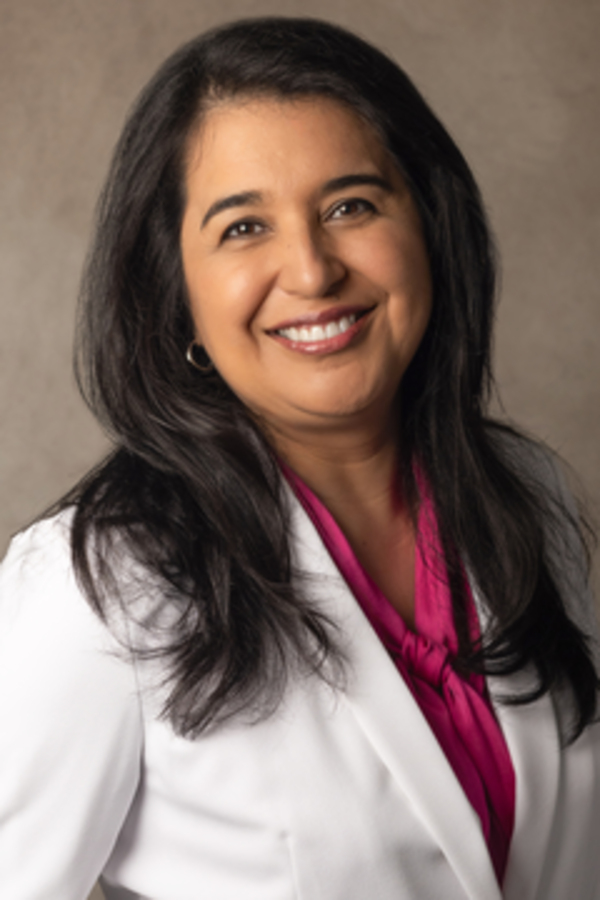 A woman with long dark hair, wearing a white blazer and a pink blouse, smiles at the camera in front of a neutral background—an advocate for raising confident kids through financial education.
