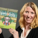 A smiling woman with blond hair holds up a children's book titled "Wooly the Bully," featuring an illustrated cow character on the cover. The background is plain and gray.