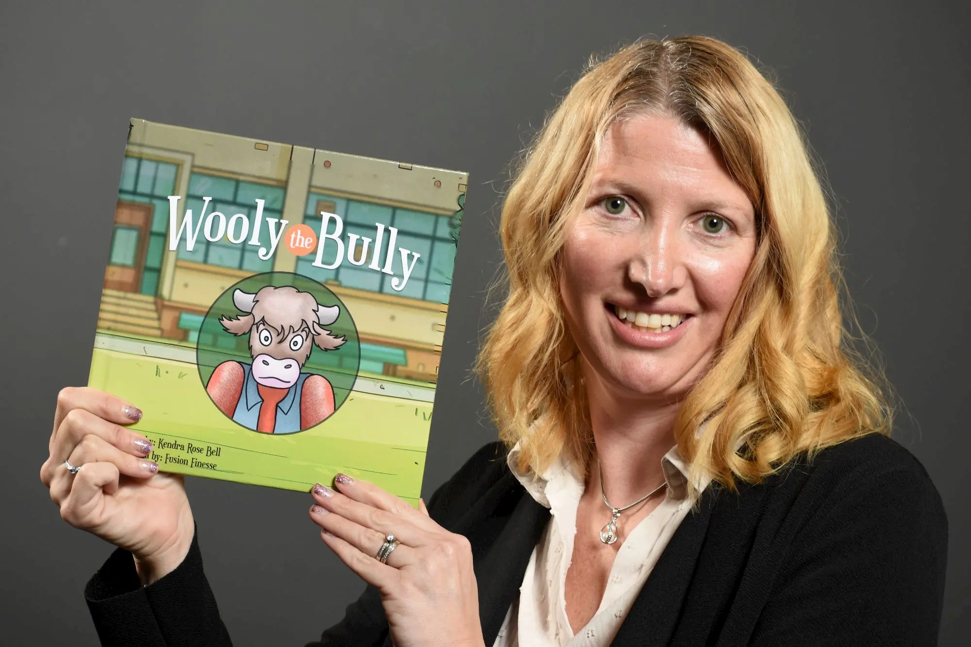 A smiling woman with blond hair holds up a children's book titled "Wooly the Bully," featuring an illustrated cow character on the cover. The background is plain and gray.
