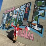A woman in a red dress and tiara stands smiling in front of a colorful mural reading “Welcome to Mobile Alabama,” holding two small flags, with her arms raised wide on a sunny day.