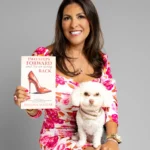 A smiling woman in a pink floral dress sits on a stool, holding a book titled "Two Steps Forward and Never Going Back." A small white dog with a collar sits on her lap.