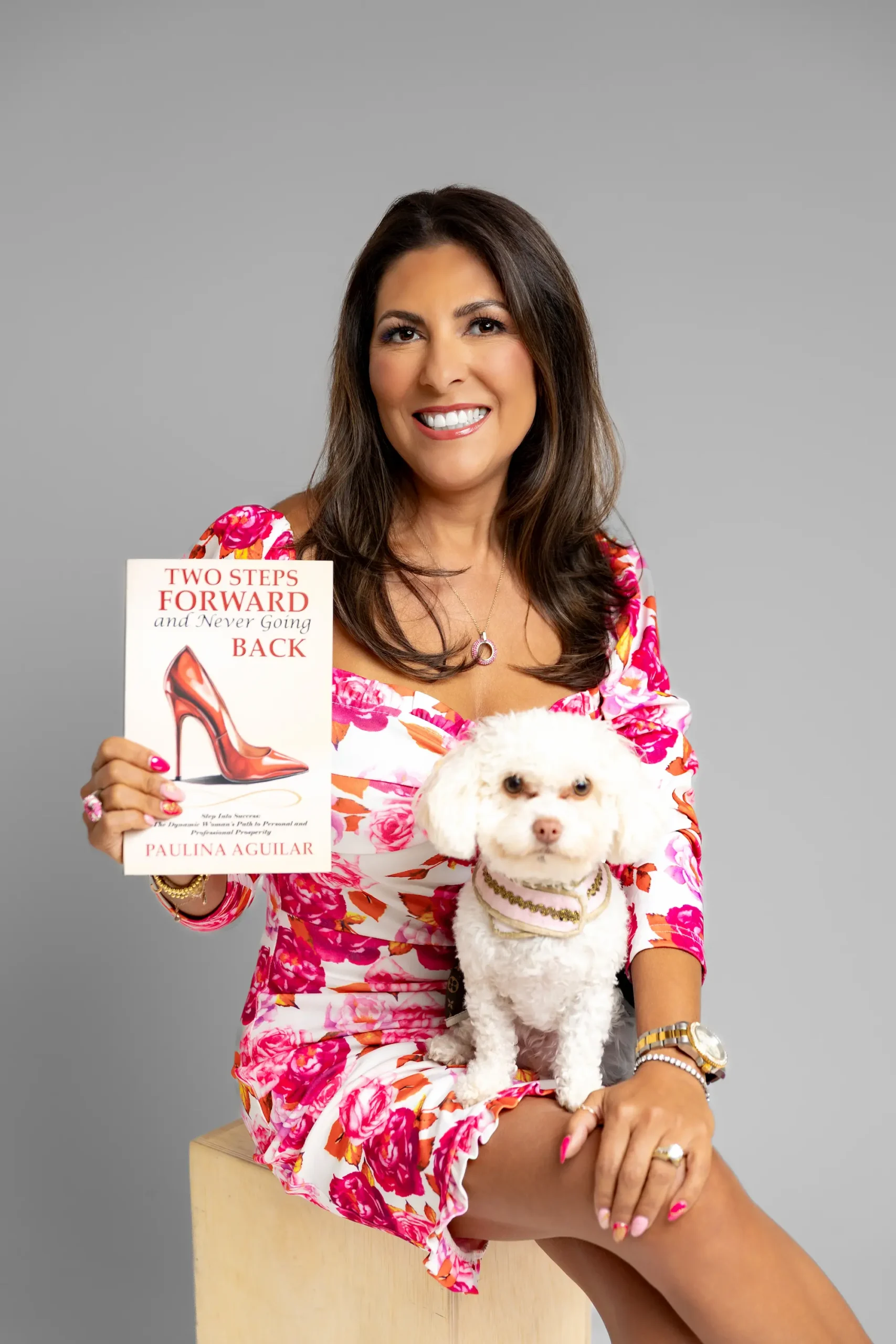 A smiling woman in a pink floral dress sits on a stool, holding a book titled "Two Steps Forward and Never Going Back." A small white dog with a collar sits on her lap.