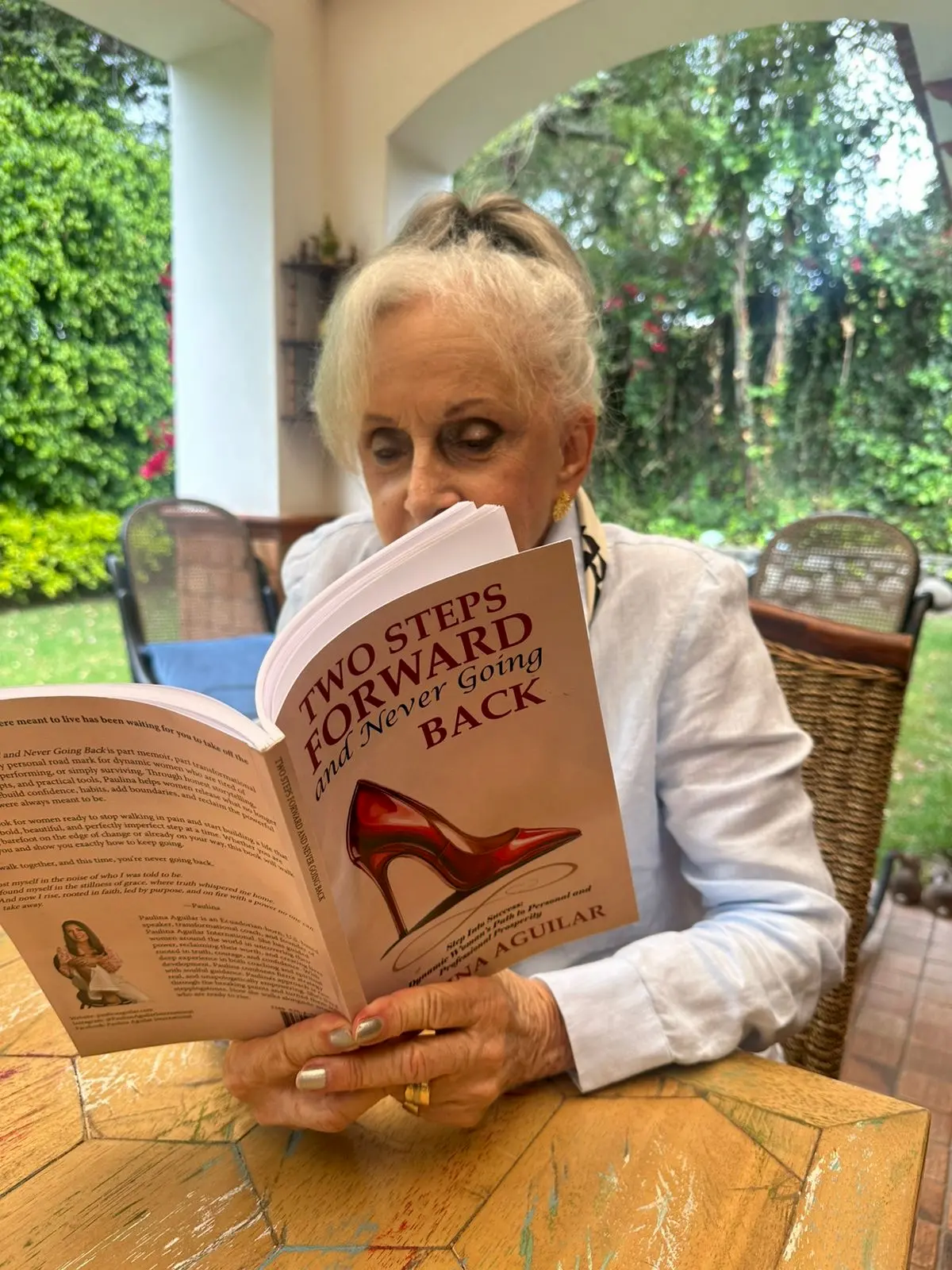 An older woman with white hair reads a book titled "Two Steps Forward and Never Going Back" by Jean Aguilar while sitting at a table outdoors on a covered patio, surrounded by greenery.