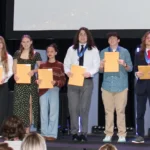 A group of eleven young people stand on a stage, smiling, and holding certificates. They appear to be receiving awards. An audience watches from the foreground. The background features a large screen.