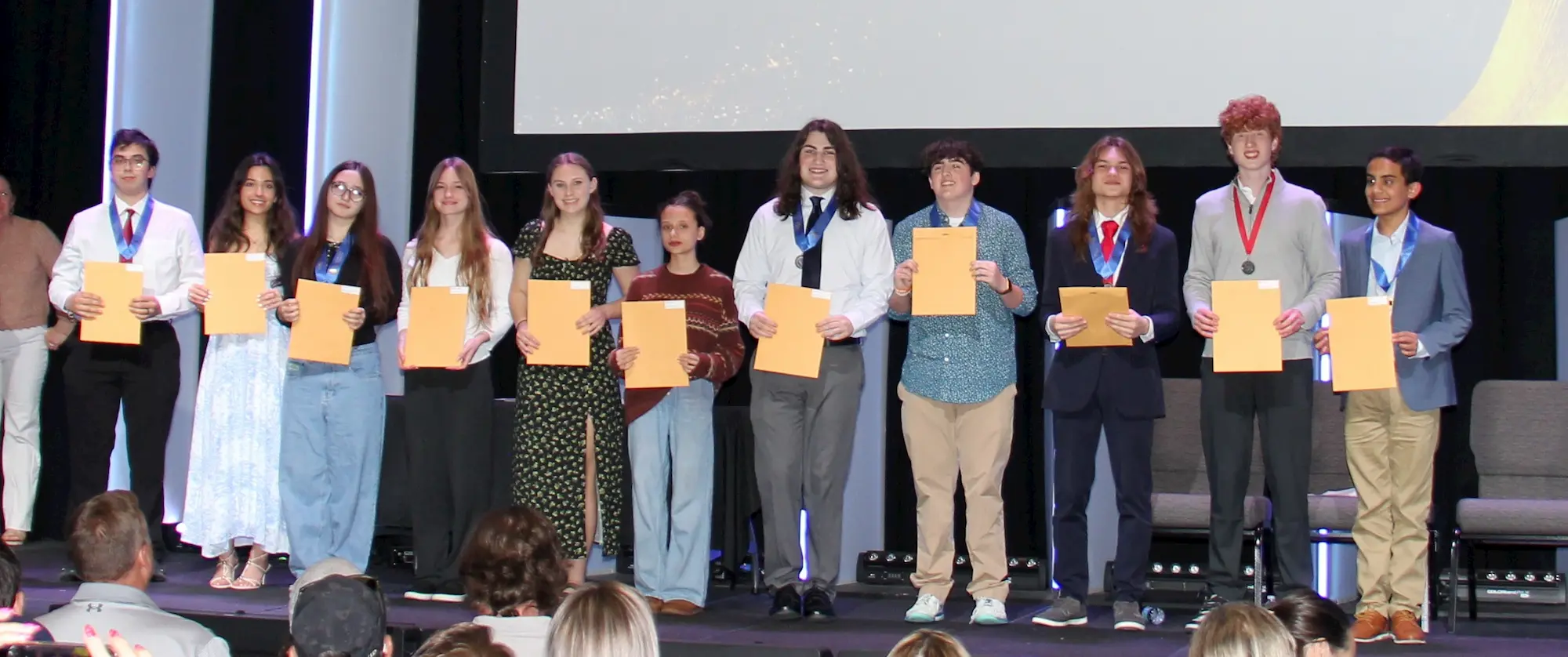 A group of eleven young people stand on a stage, smiling, and holding certificates. They appear to be receiving awards. An audience watches from the foreground. The background features a large screen.