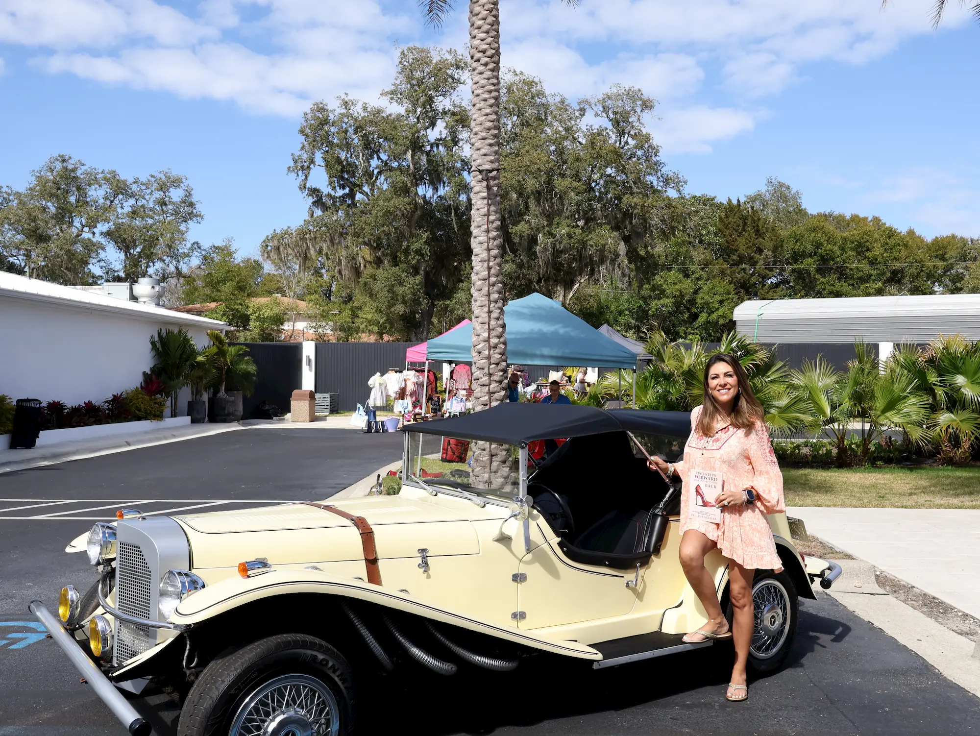 A woman in a light dress smiles while sitting on the open door of a vintage cream-colored convertible car, parked near a market with tents and palm trees in the background on a sunny day.