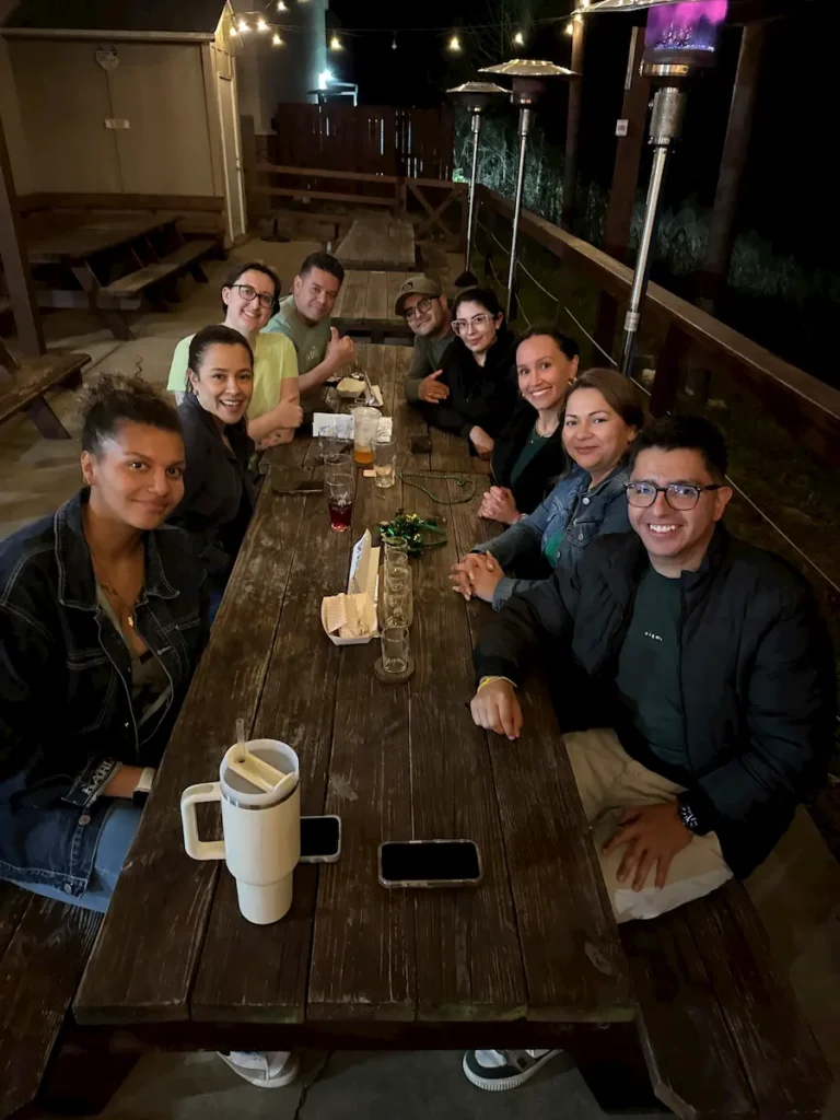 A group of nine people sits around a long wooden outdoor table at night, smiling at the camera. String lights and a patio heater are visible in the background. Cups and plates are scattered on the table.