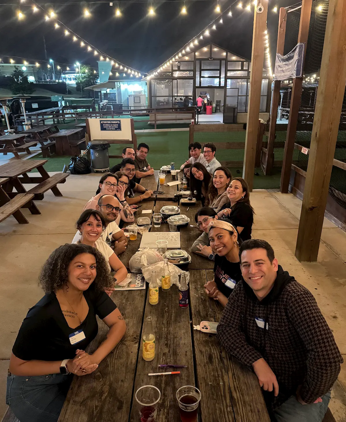 A group of people sit around a long wooden table outdoors at night, smiling and posing for a photo. String lights hang overhead, and food and drinks are on the table. Picnic tables and a fenced area are visible in the background.