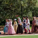 A group of people in historical colonial costumes march outdoors, some holding American flags, while onlookers sit nearby. Trees and greenery are in the background.