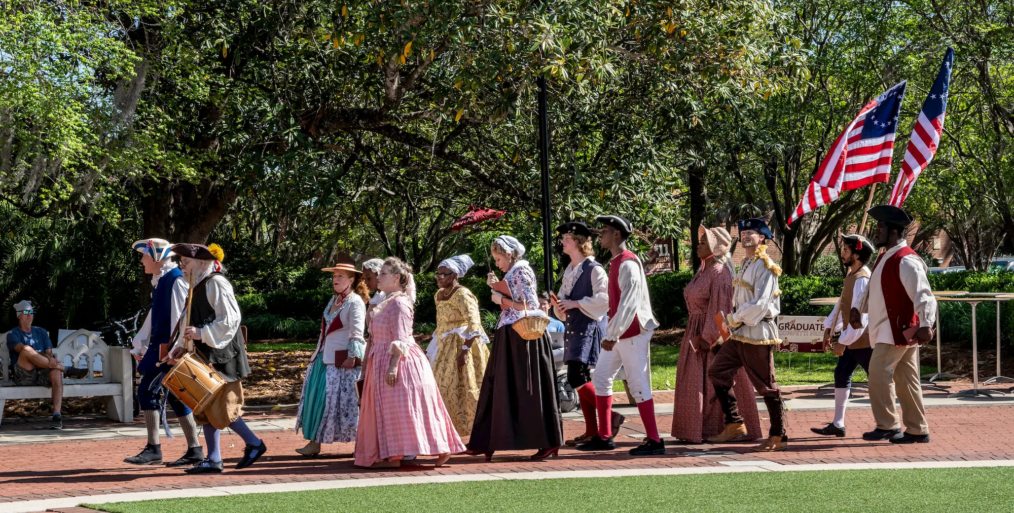 A group of people in historical colonial costumes march outdoors, some holding American flags, while onlookers sit nearby. Trees and greenery are in the background.