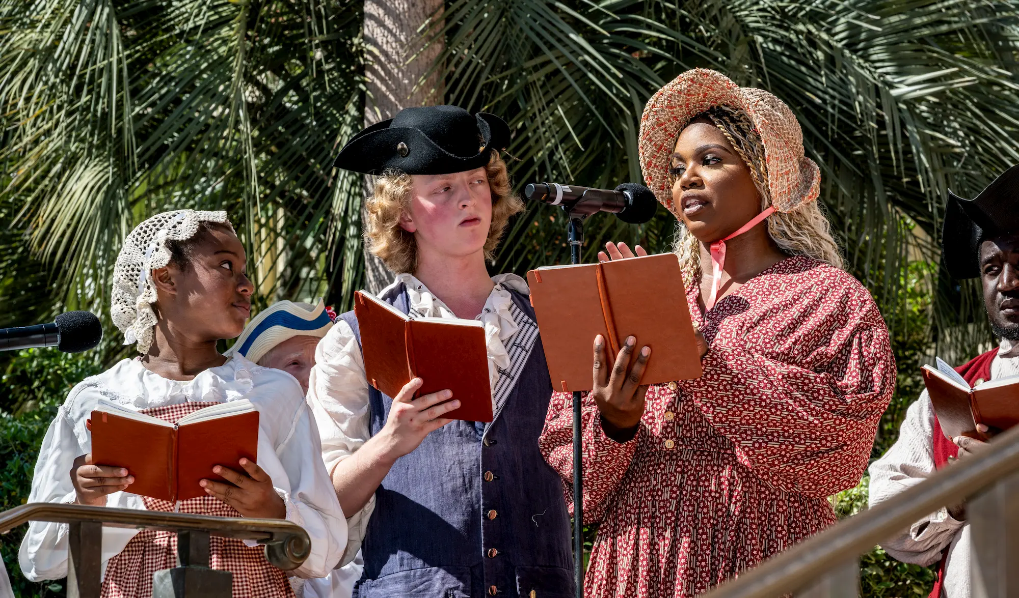 Three people dressed in historical costumes stand outdoors, holding books and reading or singing into microphones, with palm trees in the background.