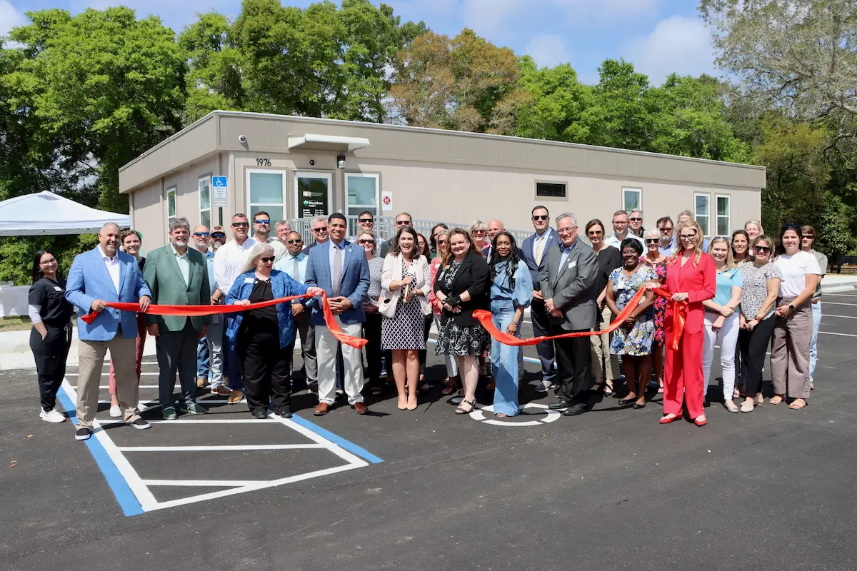 A large group of people stands in front of a small building, smiling and posing during a ribbon-cutting ceremony. Several individuals hold a long red ribbon, while others clap and celebrate on a sunny day.