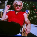 A person with short blond hair, wearing sunglasses, a red shirt, and black pants, reclines on a striped outdoor chair. They hold a cigarette and sit by a table, with greenery and red flowers in the background.