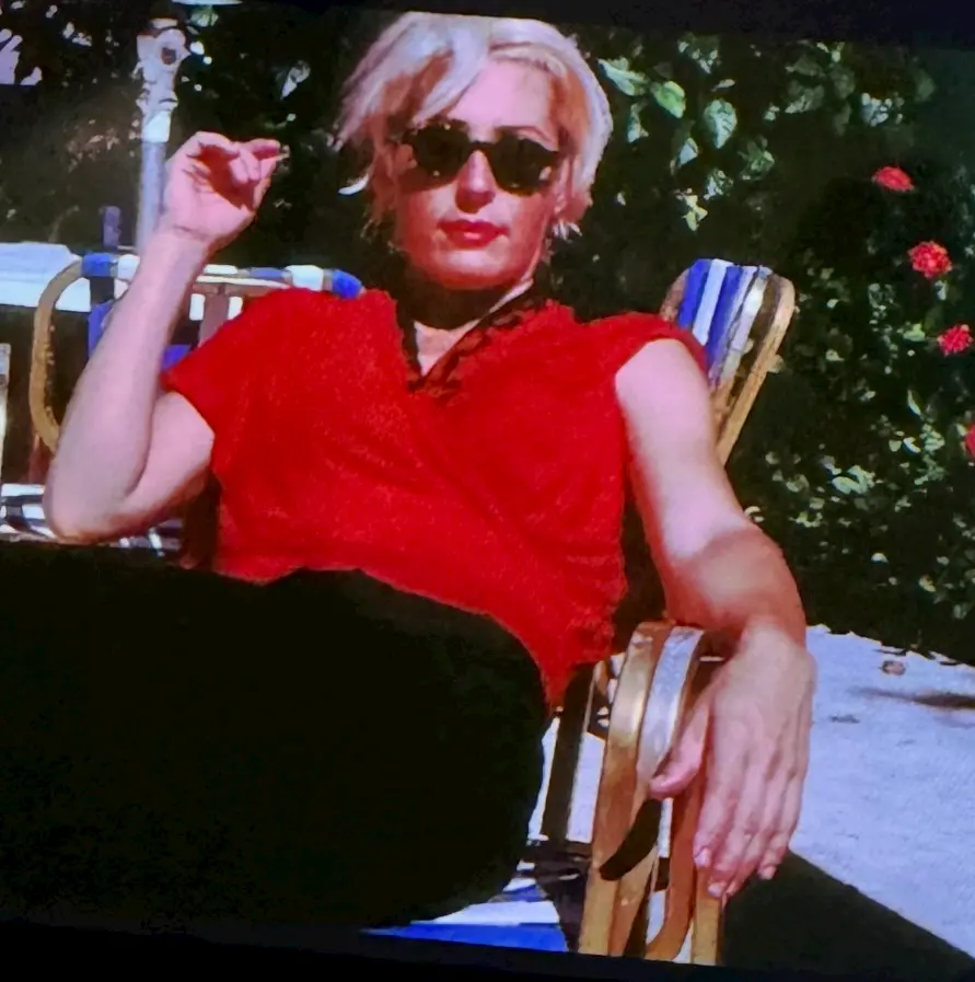 A person with short blond hair, wearing sunglasses, a red shirt, and black pants, reclines on a striped outdoor chair. They hold a cigarette and sit by a table, with greenery and red flowers in the background.