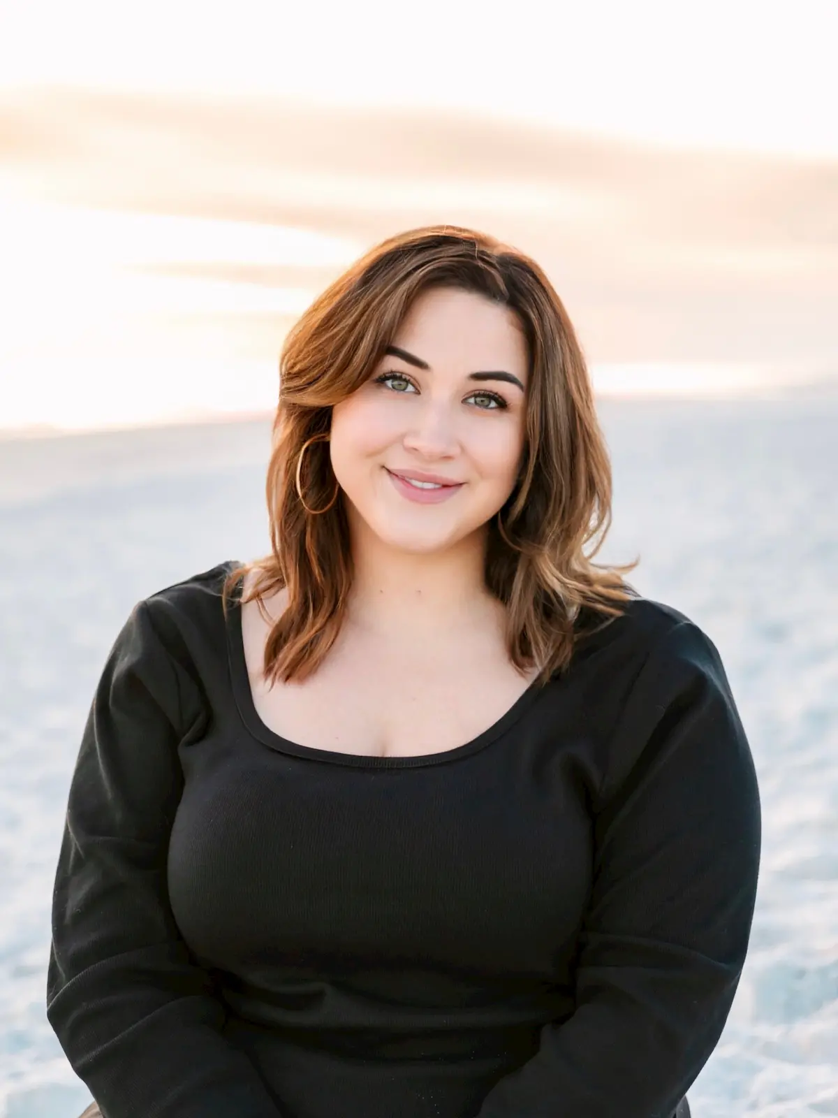 A woman with shoulder-length brown hair and hoop earrings smiles while sitting on a sandy beach, wearing a black top. The background shows a blurred shoreline and calm water at sunset.