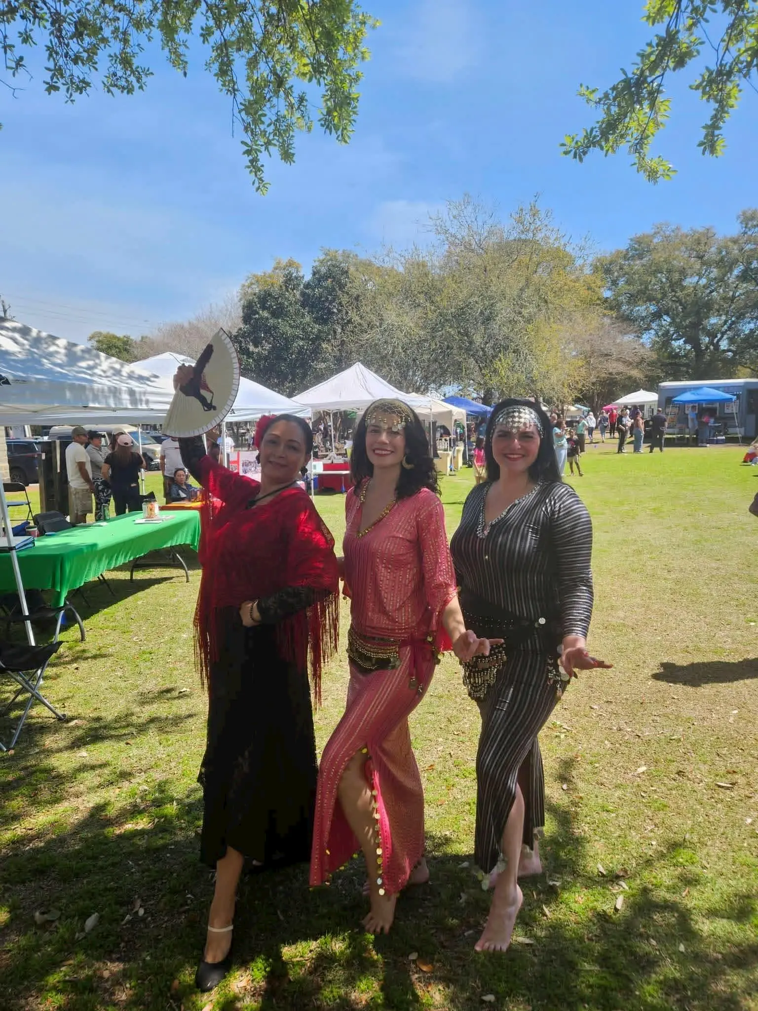 Three women in colorful, festive outfits pose and smile outdoors at a lively fair, with tents, tables, and people in the background on a sunny day, under trees with green leaves.