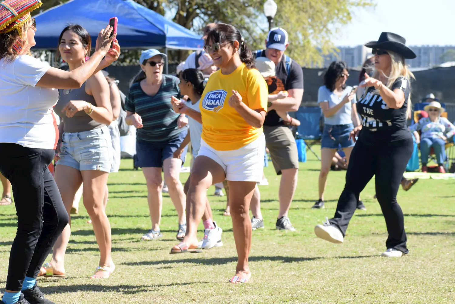 A group of people dance together on a sunny lawn at an outdoor event. They smile and laugh, wearing casual summer clothes. Some spectators relax in the background under a blue canopy and trees.