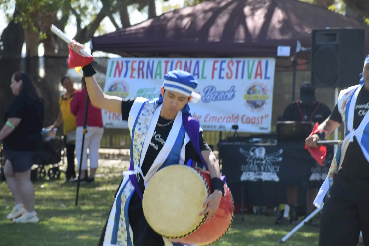 A person in traditional attire plays a drum and raises a stick at an outdoor event. A banner in the background reads "International Festival" with people and tents visible around.